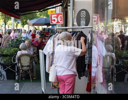 Berlino, Germania. 1 luglio 2025. 01.07.2025, Berlino. Un cittadino anziano cerca abiti usati su un appendiabiti di fronte a un negozio di tessuti usati. I vestiti sono in vendita per 5 euro ciascuno. Accanto ad esso, la gente si siede in un bar. Credito: Wolfram Steinberg/dpa credito: Wolfram Steinberg/dpa/Alamy Live News Foto Stock