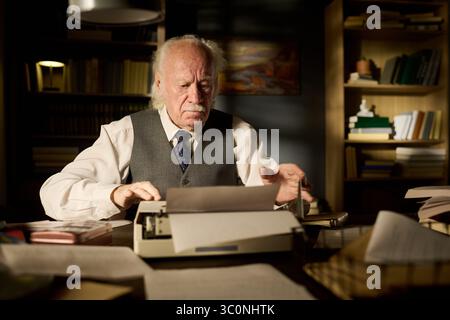 Uomo caucasico seduto alla scrivania usando una macchina da scrivere, concentrandosi sulla scrittura circondato da pile di carte e libri in ufficio o in studio con scaffali sullo sfondo Foto Stock