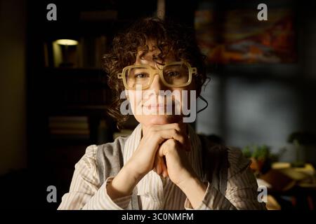 Ritratto di una donna caucasica di mezza età con capelli ricci e occhiali che sorridono delicatamente mentre si riposa il mento sulle mani, guardando direttamente la fotocamera in un ambiente interno con luce soffusa Foto Stock