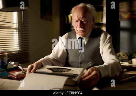 Uomo caucasico seduto alla scrivania usando una macchina da scrivere, concentrandosi sulla scrittura di documenti, indossando camicia e gilet formali, circondato da libri e documenti in ufficio Foto Stock