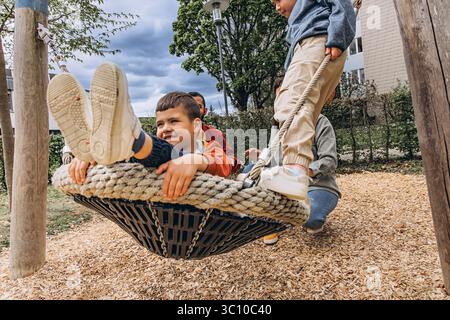 Famiglia felice con due figli che giocano insieme su un grande altalena di corda al parco giochi. I genitori sorridono, legano e trascorrono del tempo di qualità con i bambini all'aperto Foto Stock