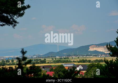 Case suburbane e terreni agricoli con una cava e un camino industriale in lontananza, visti da un bordo della foresta in una giornata estiva Foto Stock