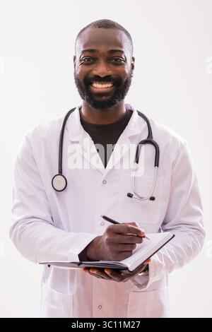 Ritratto di allegro nero maschio paramedico in uniforme medica che sta scrivendo note in organizzatore personale. Studio girato su sfondo grigio. Foto Stock