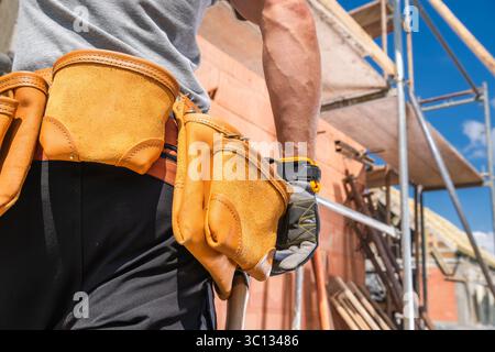 Un operaio edile con una camicia grigia e una cintura portautensili è pronto in un cantiere. Ponteggi e materiali sono visibili sotto un cielo azzurro. Foto Stock