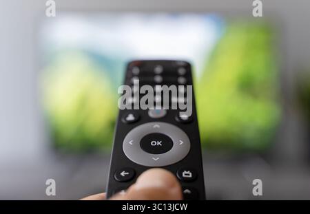 Una mano che tiene in mano il telecomando commuta i canali di un televisore con uno schermo sfocato sullo sfondo. Le riprese simboleggiano il moderno consumo dei media, il sovraccarico di contenuti, l'affaticamento delle scelte digitali e la costante ricerca di informazioni coinvolgenti. Ideale per temi riguardanti le abitudini multimediali, l'intrattenimento, l'età dell'informazione e la dipendenza dallo schermo. Foto Stock