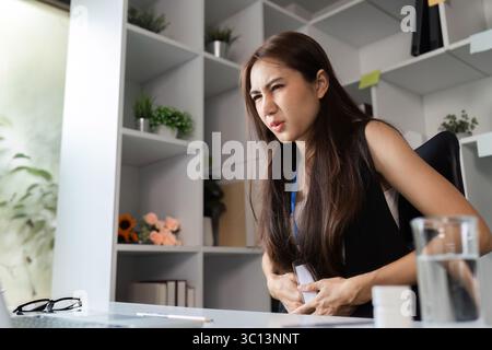 Donna d'affari che si sente male al lavoro, seduta alla scrivania con espressione dolorosa in un moderno ambiente d'ufficio Foto Stock