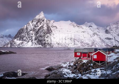 Vista delle vivaci cabine rosse annidate sulla costa rocciosa e innevata, di fronte a una maestosa montagna innevata sotto un cielo spettacolare, Hamnøy, Norvegia. Foto Stock