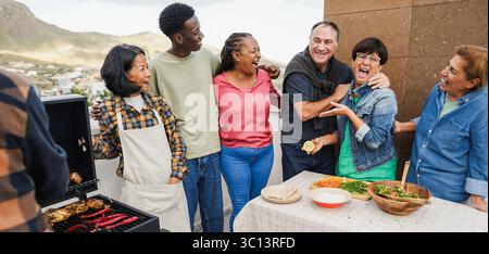 Persone multigenerazionali che fanno barbecue sul tetto di casa - amici multirazziali che si divertono a mangiare e cucinare insieme durante il fine settimana Foto Stock