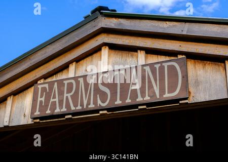 Un cartello in legno con la scritta “FARMSTAND” montato sul timpano di un rustico edificio in legno, adagiato contro un cielo blu. Foto Stock