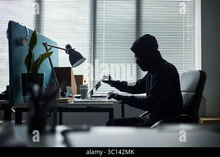 Giovane uomo caucasico che indossa cappuccio nero, maschera facciale e guanti seduto alla scrivania utilizzando il computer portatile in un ufficio moderno, sembra accedere al computer in segreto, rubare dati aziendali o rompere nel sistema Foto Stock