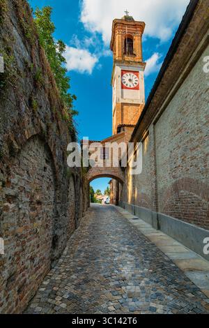 Stretta strada acciottolata tra mura in pietra, che conduce a un campanile con orologio a Guarene, Italia. Foto Stock