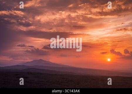 La vista aerea di un tramonto infuocato dipinge l'orizzonte con arance e viola vibranti, gettando ombre sulle montagne lontane, posizione sconosciuta. Foto Stock
