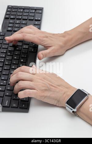 Donna lavoratrice che scrive su una tastiera nera e indossa uno smartwatch al polso sinistro Foto Stock