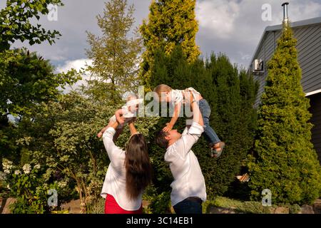 Madre e padre sollevano contemporaneamente il loro figlio e la loro bambina contro gli alti sempreverdi, catturando risate energiche e il lavoro di squadra in Golden ev Foto Stock