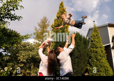 Nella luce della sera dorata, il padre getta in alto il figlio ridendo, mentre la madre solleva il bambino, giocherellando in famiglia incorniciato da alti sempreverdi e backya Foto Stock