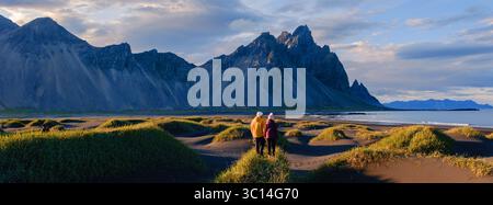 Due avventurosi escursionisti si stagliano su una spiaggia sabbiosa tra lussureggianti prati verdi, ammirando la splendida montagna Vestrahorn. Le tonalità dorate dell'alba illuminano il paesaggio aspro di Stokksnes, Islanda. Foto Stock