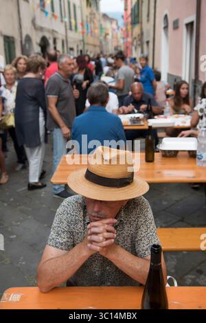 Borgomaro, un villaggio medievale italiano di montagna, organizza il suo festival annuale di conchiglie e cozze. I tavoli riempiono la strada principale, un uomo locale aspetta con una bottiglia di vino per gli amici. Borgomaro, regione Liguria, Italia, Europa. Luglio 2025 HOMER SYKES Foto Stock