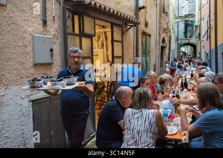 Italia festival gastronomico regionale Europa. Borgomaro, un villaggio medievale italiano, organizza il suo festival annuale di conchiglie e cozze. I tavoli riempiono una stretta strada laterale, i residenti locali e gli amici si godono un pasto comune. Un uomo del posto con un vassoio con quattro ciotole di cozze cammina fino alla sua tavola. Borgomaro, regione Liguria, Italia. Luglio 2025 HOMER SYKES Foto Stock