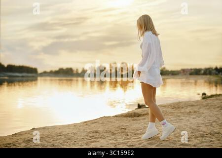 Donna che cammina sulla spiaggia sabbiosa al tramonto in acque calme in un tranquillo ambiente naturale Foto Stock
