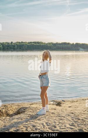 Una giovane donna si erge sulla spiaggia sabbiosa di un lago calmo, ammirando il tramonto. La sua posa rilassata e il suo outfit casual creano un'atmosfera tranquilla e raffinata Foto Stock