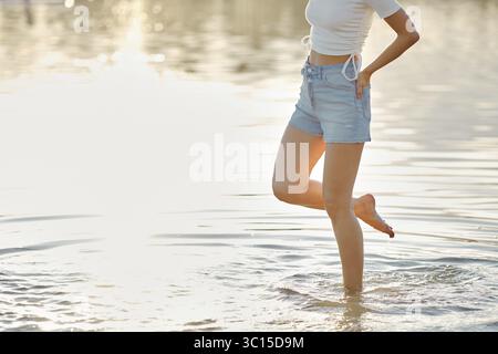 Una donna irriconoscibile cammina con attenzione nell'acqua limpida, godendosi il calore del sole sulla pelle. Indossa un top leggero e un paio di pantaloncini in denim Foto Stock