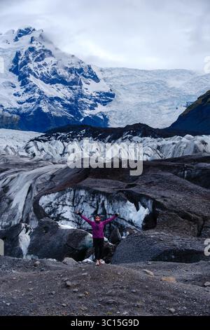 Un viaggiatore si erge sul terreno accidentato del ghiacciaio Svinafellsjokull, meravigliandosi davanti alle formazioni ghiacciate mozzafiato e alle torreggianti vette innevate. Questa splendida posizione incarna la selvaggia bellezza delle ghiacciaie. Foto Stock