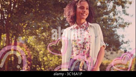 Donna sorridente che indossa camicia e fascia floreale pastello nel parco illuminato dal sole, con sovrapposizioni arcobaleno a cuore Foto Stock