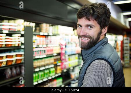 Un acquirente maschile che si trova nella corsia del supermercato e indossa un giubbotto trapuntato e un maglione tra gli scaffali riforniti Foto Stock