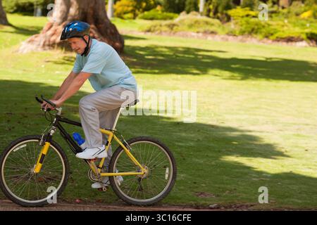 Uomo anziano in bicicletta gialla con casco blu, bottiglia d'acqua sul telaio nel parco, spazio fotocopie Foto Stock