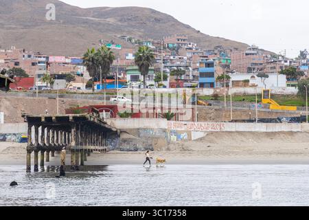 Una spiaggia di Chancay con un molo e alcuni edifici sullo sfondo. Una donna e un cane camminano sulla spiaggia Foto Stock