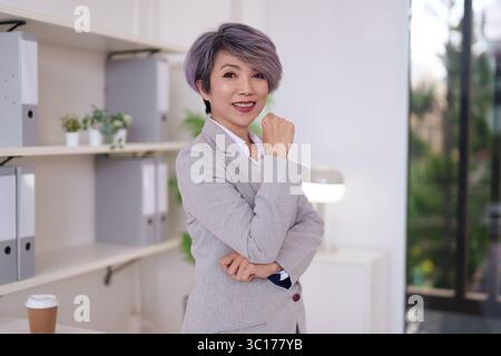 Donna adulta asiatica professionista di affari sorride con fiducia in piedi in un ambiente d'ufficio moderno successo di carriera Foto Stock