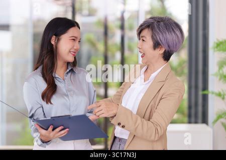Due donne asiatiche sorridenti che consultano documenti di lavoro in un ambiente luminoso e moderno. Condividono le idee collaborano al tutorship dei progetti. Foto Stock