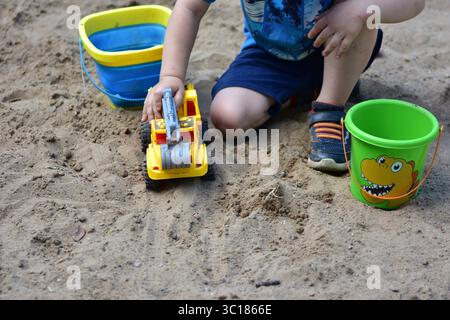Bambino anonimo che gioca nella sandbox con un camion giocattolo giallo e due secchi, circondato da sabbia asciutta in una giornata di sole, catturato durante i primi anni di chi Foto Stock