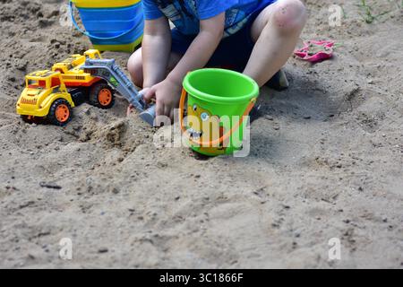 Ragazzo che gioca nella sandbox con un camion giocattolo giallo e due secchi, circondato da sabbia secca e erba in una giornata di sole, catturato durante la prima infanzia p Foto Stock