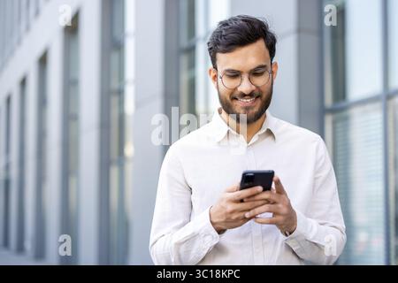 Un uomo sorridente con gli occhiali guarda il suo telefono mentre si trova di fronte a un ufficio. Foto Stock