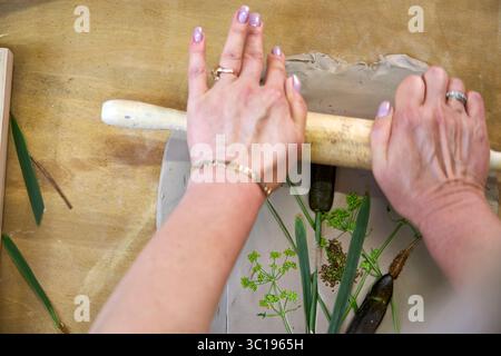 Premendo fiori e foglie in argilla con un mattarello per creare un'impronta botanica per il rilievo gesso. Mani appiattimento piante in fai da te passo per passo t Foto Stock