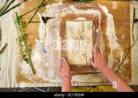 Mani che tengono il rilievo in gesso botanico finito in una cornice di legno. Opere d'arte floreale fai da te con stampe di fiori e foglie, fase finale del decoro fatto a mano Foto Stock