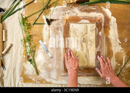 Mani che tengono il rilievo in gesso botanico finito in una cornice di legno. Opere d'arte floreale fai da te con stampe di fiori e foglie, fase finale del decoro fatto a mano Foto Stock