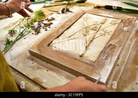 Mani che tengono il rilievo in gesso botanico finito in una cornice di legno. Opere d'arte floreale fai da te con stampe di fiori e foglie, fase finale del decoro fatto a mano Foto Stock