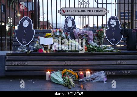 Broad Street, Birmingham, 22 luglio 2025: I fan di Ozzy Osbourne sono venuti al Black Sabbath Bridge di Birmingham in Broad Street per rendere omaggio a Ozzy Osbourne, morto all'età di 76 anni. Fiori, biglietti e candele sono stati lasciati sulla panchina e alcune persone hanno bevuto qualcosa e li hanno condivisi con l'immagine di Ozzy sulla panchina. Crediti: British News and Media/Alamy Live News Foto Stock