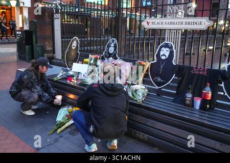 Broad Street, Birmingham, 22 luglio 2025: I fan di Ozzy Osbourne sono venuti al Black Sabbath Bridge di Birmingham in Broad Street per rendere omaggio a Ozzy Osbourne, morto all'età di 76 anni. Fiori, biglietti e candele sono stati lasciati sulla panchina e alcune persone hanno bevuto qualcosa e li hanno condivisi con l'immagine di Ozzy sulla panchina. Crediti: British News and Media/Alamy Live News Foto Stock