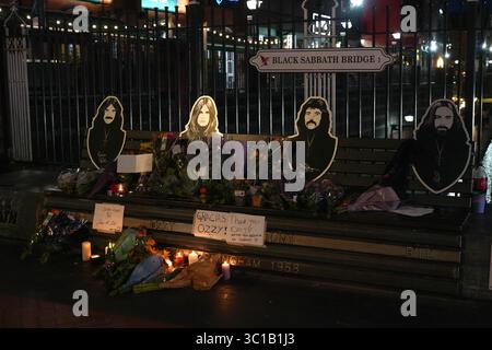 Broad Street, Birmingham, 22 luglio 2025: I fan di Ozzy Osbourne sono venuti al Black Sabbath Bridge di Birmingham in Broad Street per rendere omaggio a Ozzy Osbourne, morto all'età di 76 anni. Fiori, biglietti e candele sono stati lasciati sulla panchina e alcune persone hanno bevuto qualcosa e li hanno condivisi con l'immagine di Ozzy sulla panchina. Crediti: British News and Media/Alamy Live News Foto Stock