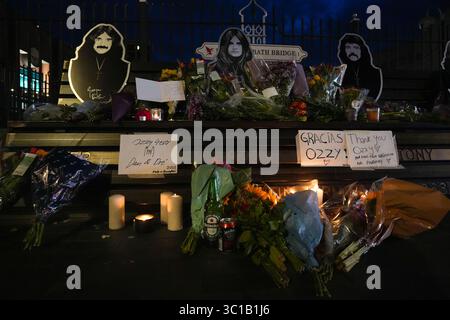 Broad Street, Birmingham, 22 luglio 2025: I fan di Ozzy Osbourne sono venuti al Black Sabbath Bridge di Birmingham in Broad Street per rendere omaggio a Ozzy Osbourne, morto all'età di 76 anni. Fiori, biglietti e candele sono stati lasciati sulla panchina e alcune persone hanno bevuto qualcosa e li hanno condivisi con l'immagine di Ozzy sulla panchina. Crediti: British News and Media/Alamy Live News Foto Stock