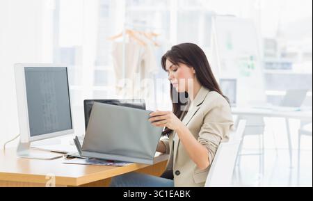 Donna che lavora su un computer portatile con monitor esterno e appendiabiti dietro mentre è seduta alla scrivania dell'ufficio Foto Stock