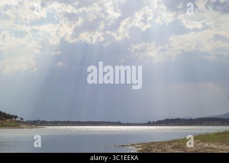 Vista panoramica dei raggi del sole che filtrano attraverso spesse nuvole, che proiettano la luce su un lago calmo e una costa lontana. Atmosfera tranquilla e serena, perfetta Foto Stock