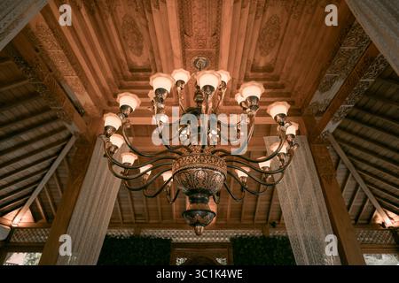 Un magnifico lampadario in stile Giavanese tradizionale appeso ad un intricato soffitto in legno intagliato. Vista ad angolo basso del lusso e degli interni classici Foto Stock