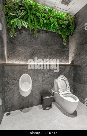 Bagno moderno con WC e orinatoio contro pareti in marmo scuro, con design biofilo con un lussureggiante soffitto di piante artificiali verdi Foto Stock