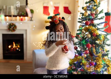 Ragazza afro-americana sorridente e in possesso di un ornamento rosso in baule nel soggiorno vicino all'albero di Natale Foto Stock