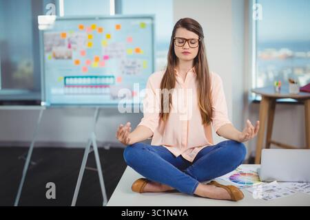 Donna che meditava con le mani appoggiate sulle ginocchia sulla scrivania in un ufficio moderno con note sulla lavagna bianca Foto Stock