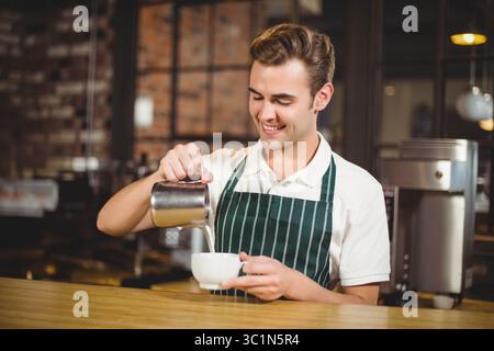 Barista maschio che versa il latte montato dalla caraffa metallica nella tazza accanto alla macchina dietro il bancone del caffè Foto Stock